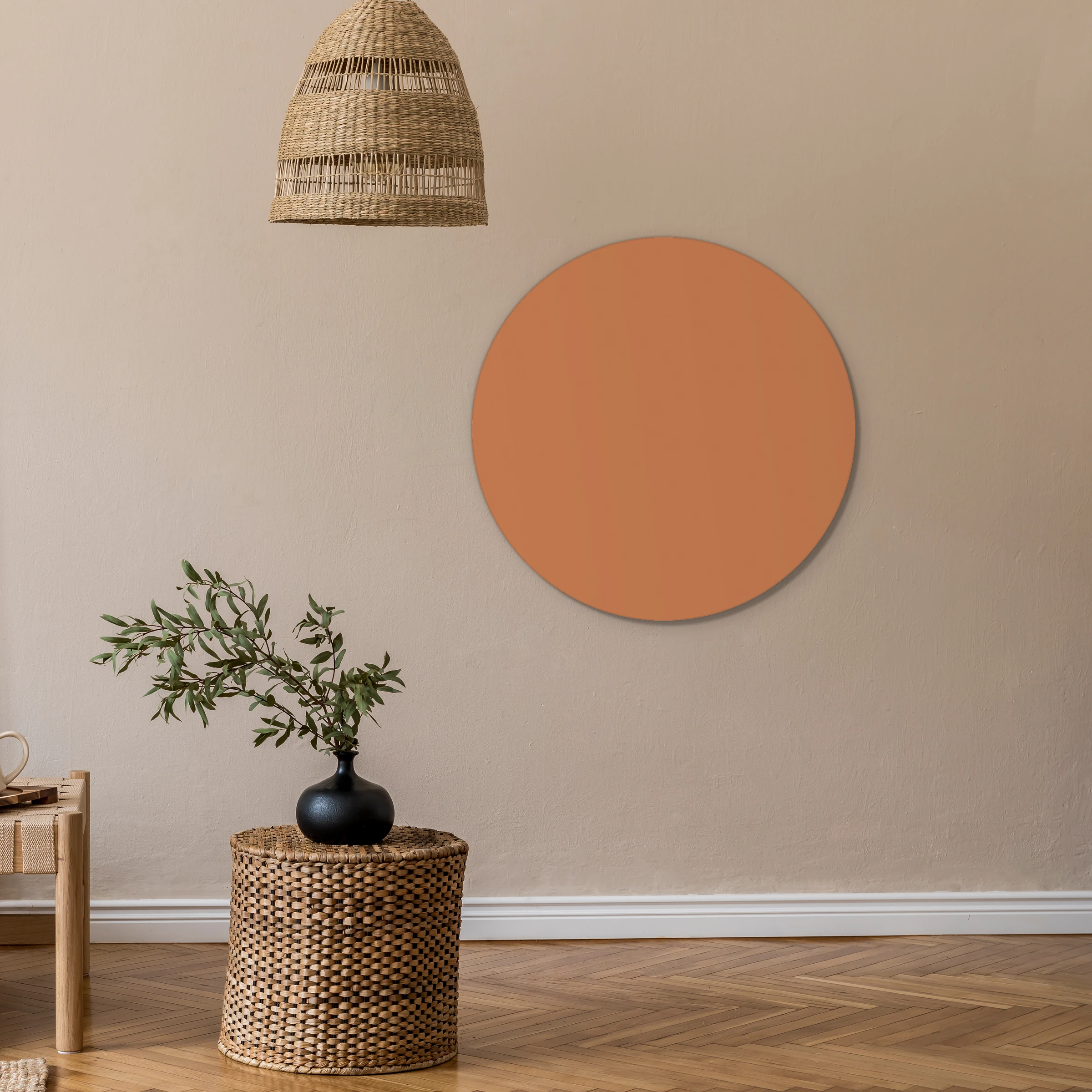 Muurcirkel zandbruin aan een beige wand onder een rieten hanglamp, met een olijftak op een rieten krukje